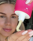 Person holding a pink bottle with a white pump, dispensing a white substance onto their face.