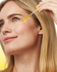 A close-up of a woman with blonde hair applying a golden-yellow serum or oil dropper to her cheek, set against a soft yellow gradient background.