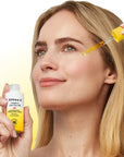 A woman applies Derma-E Vitamin Glam Face Oil with a dropper to her face while holding the white and yellow bottle in the other hand, set against a bright yellow gradient background.