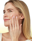 A close-up of a woman with blonde hair applying a cream product to her cheek, shown against a clean white and yellow gradient background.