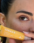 A close-up of an holding a yellow tube of DERMA E Vitamin C No Dark Circles Perfecting Eye Cream.