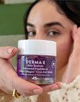 Close-up of a hand holding a Derma E Skin Restore Cryo-Gel Mask jar in the foreground while another hand applies product to the face in the background.