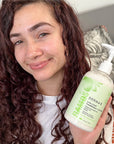 The image shows a women holding a bottle of DERMA‑E 2‑in‑1 Curl Conditioner + Co‑Wash in front of their curly hair. The focus is on the product bottle and defined curls, highlighting the product's use in real hair‑care routines.