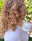 A women holds a jar of DERMA‑E Curl Repair Deep Treatment next to their defined, bouncy curls. The focus is on the product jar and the healthy, hydrated curls, illustrating real‑life results after using the treatment.