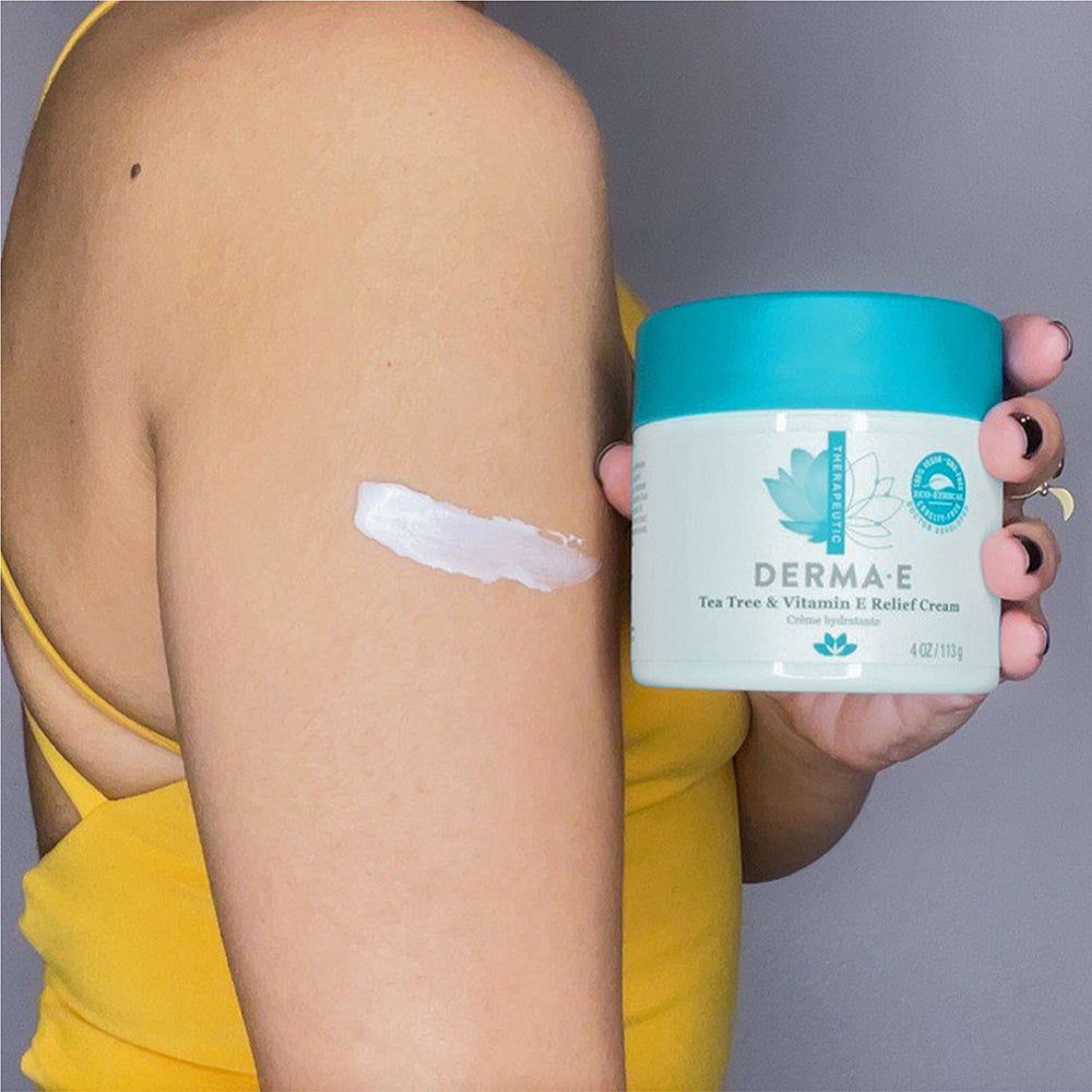 Woman holding Derma E's Tea Tree and Vitamin E Relief Cream jar with smear on her arm.