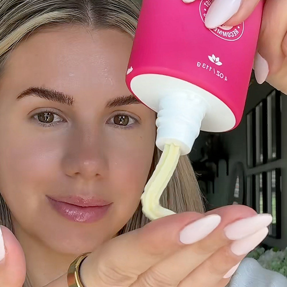 Person holding a pink bottle with a white pump, dispensing a white substance onto their face.