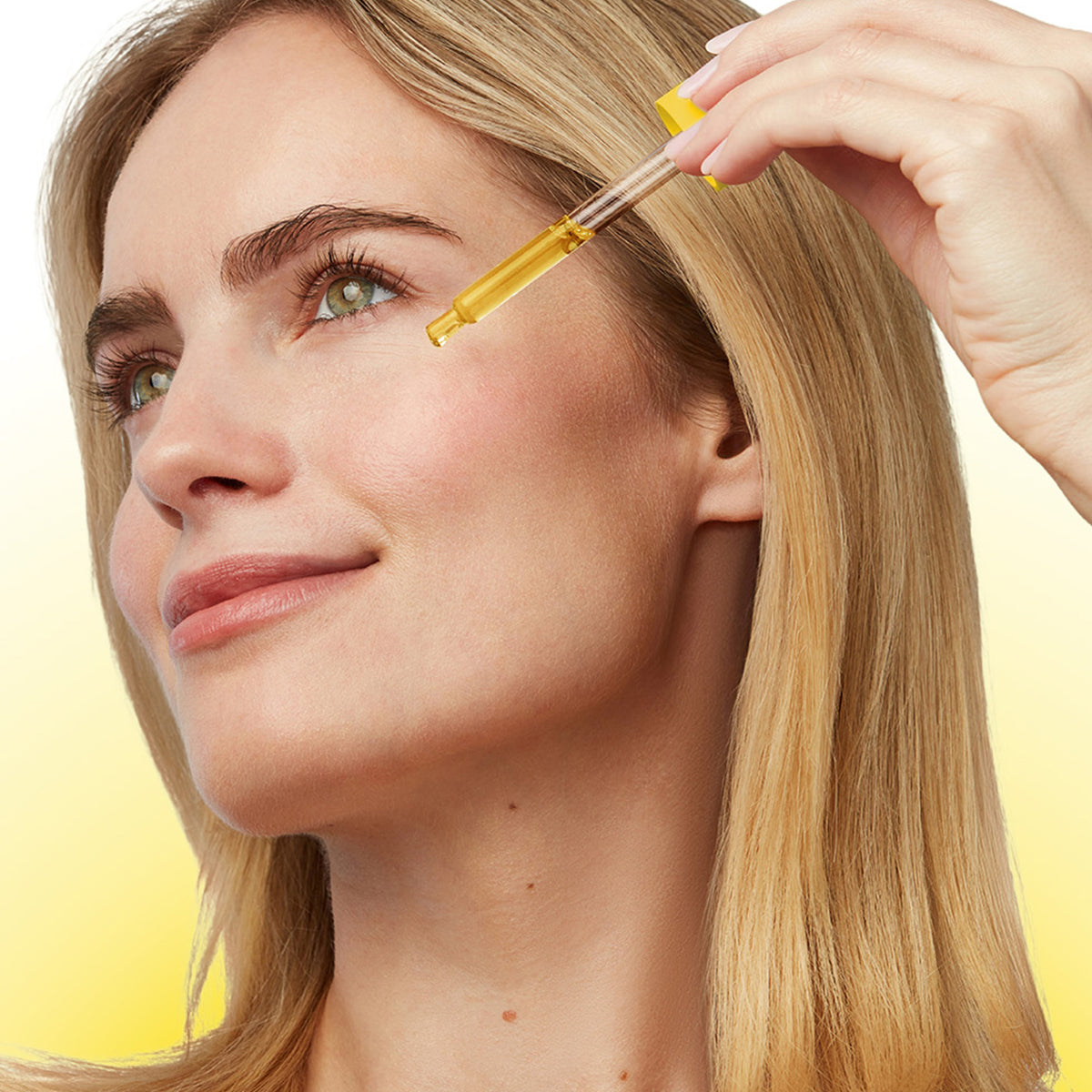 A close-up of a woman with blonde hair applying a golden-yellow serum or oil dropper to her cheek, set against a soft yellow gradient background.