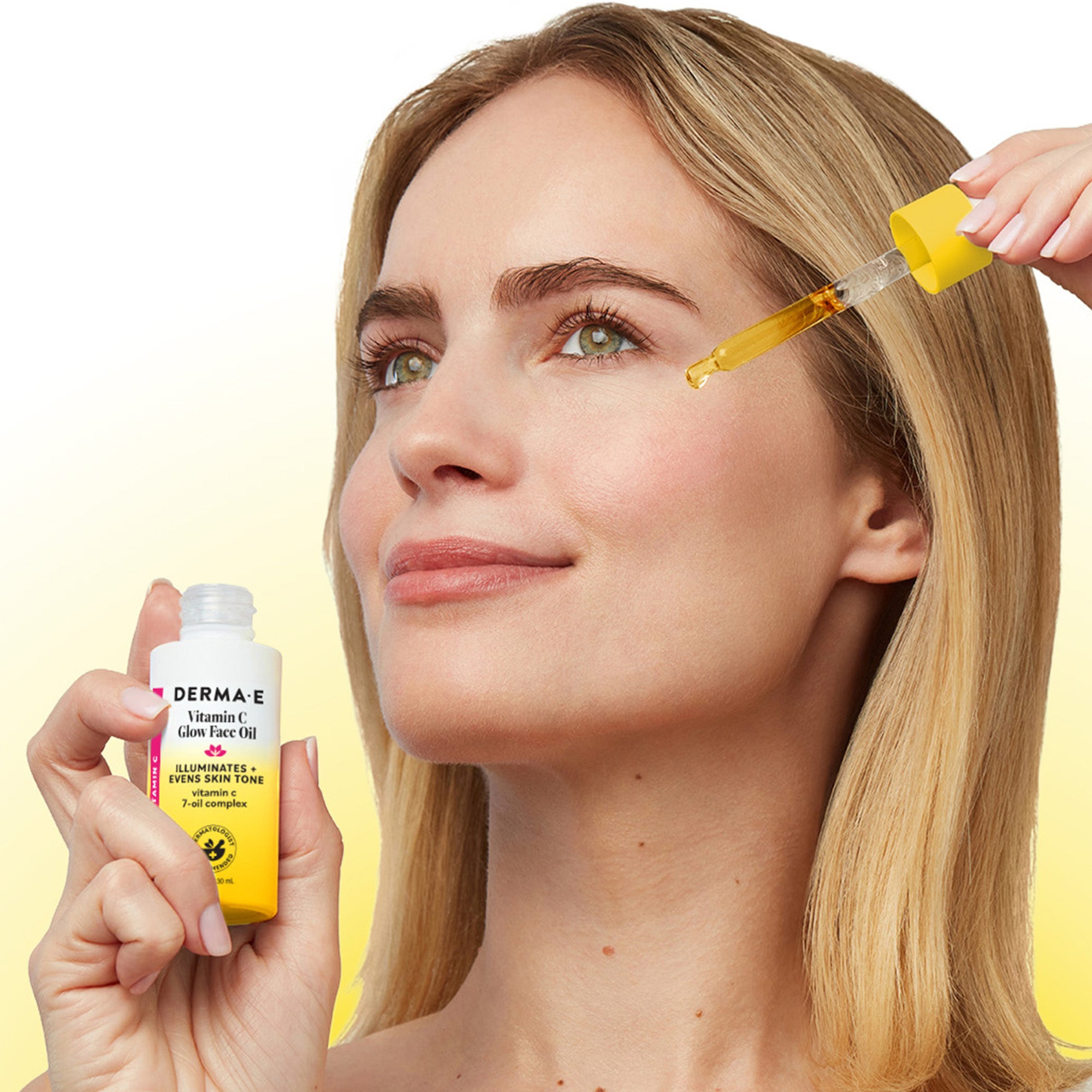 A woman applies Derma-E Vitamin Glam Face Oil with a dropper to her face while holding the white and yellow bottle in the other hand, set against a bright yellow gradient background.