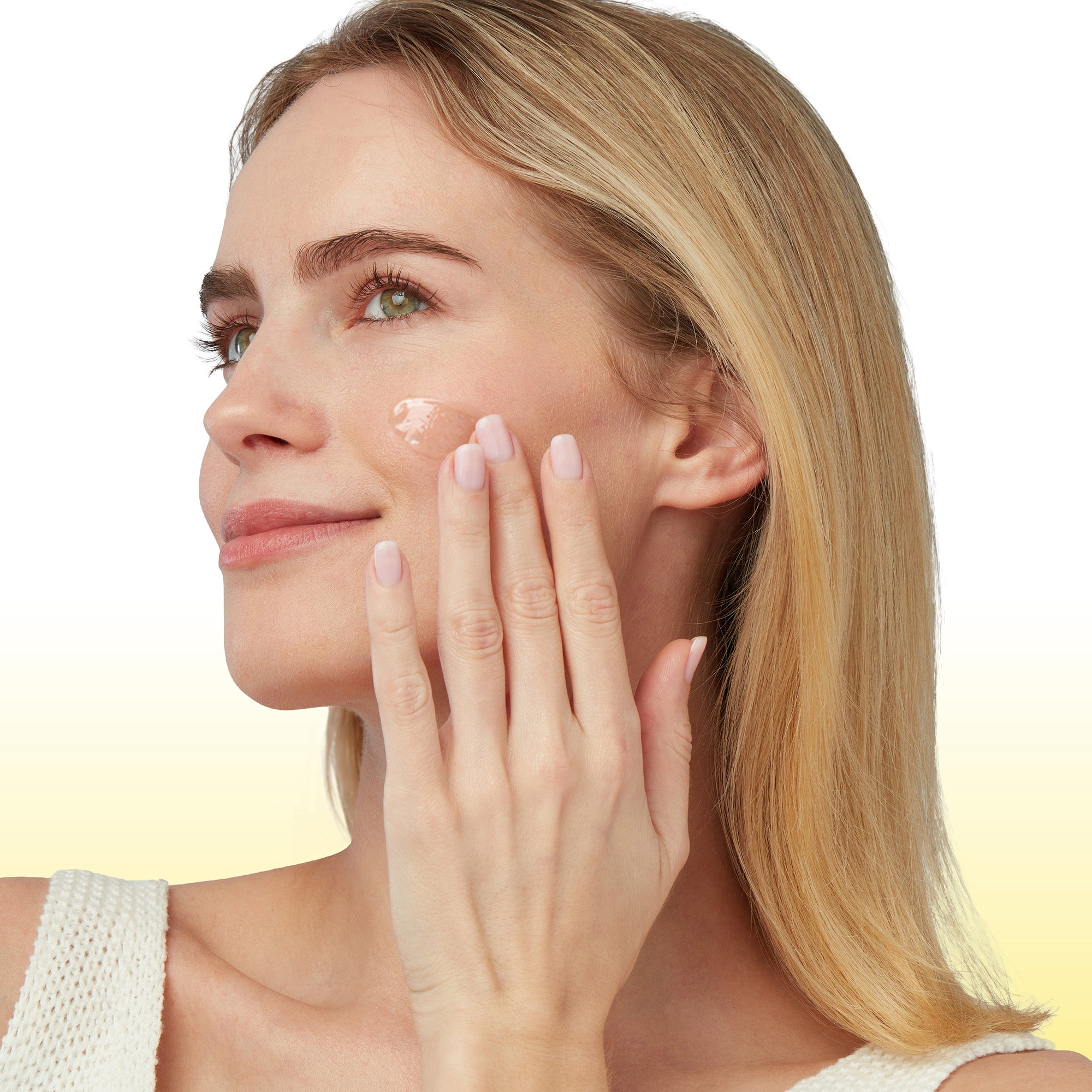 A close-up of a woman with blonde hair applying a cream product to her cheek, shown against a clean white and yellow gradient background.
