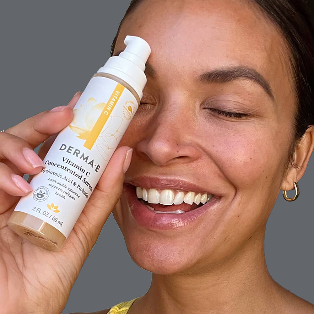 Smiling woman holding Derma E Vitamin C Serum bottle near her face, highlighting the product&#39;s packaging and the natural glow of her skin.