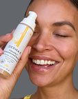 Smiling woman holding Derma E Vitamin C Serum bottle near her face, highlighting the product's packaging and the natural glow of her skin.